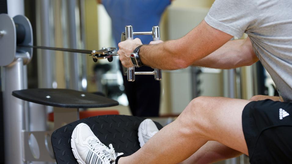 Mann trainiert im Fitnessstudio mit einem Kabelzuggerät, ein anderer Mensch im Hintergrund führt eine Übung aus.